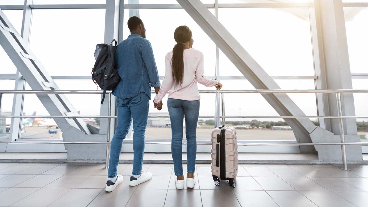 A couple holds hands while looking at an airplane taking off through an airport window