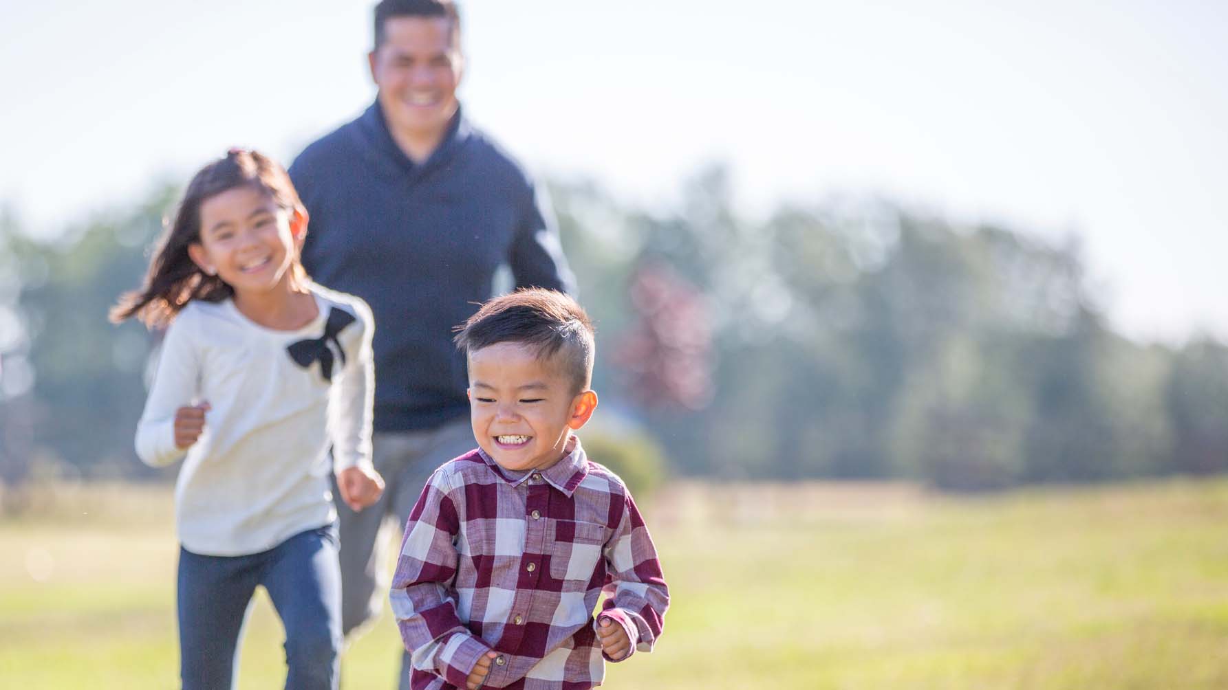 A man and his two young children run in a field while laughing