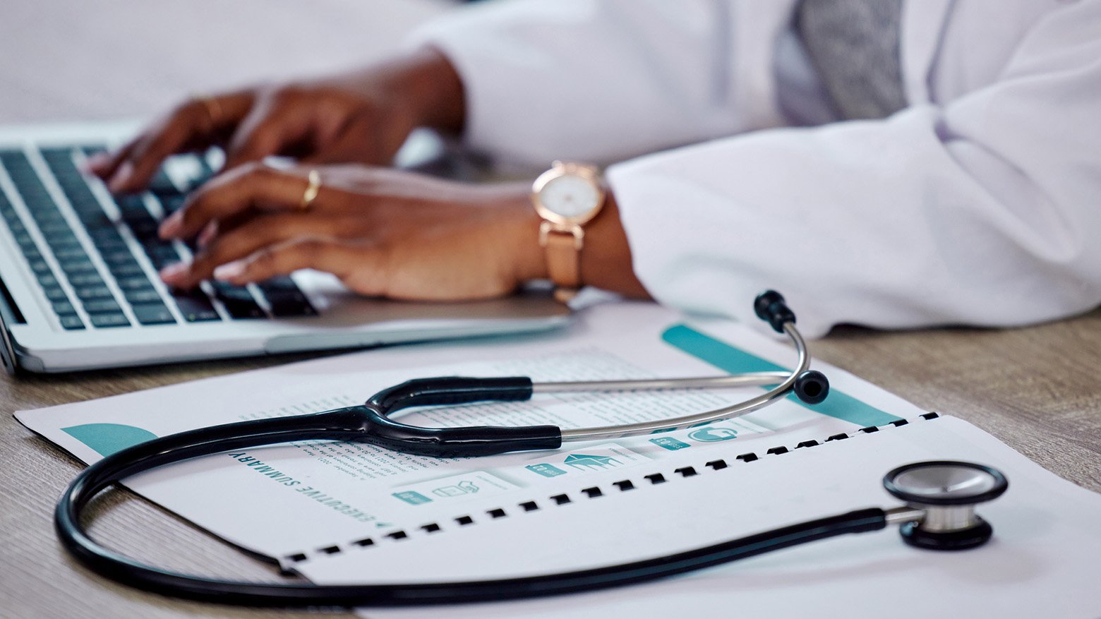 A doctor's hands typing on a laptop next to a stethoscope