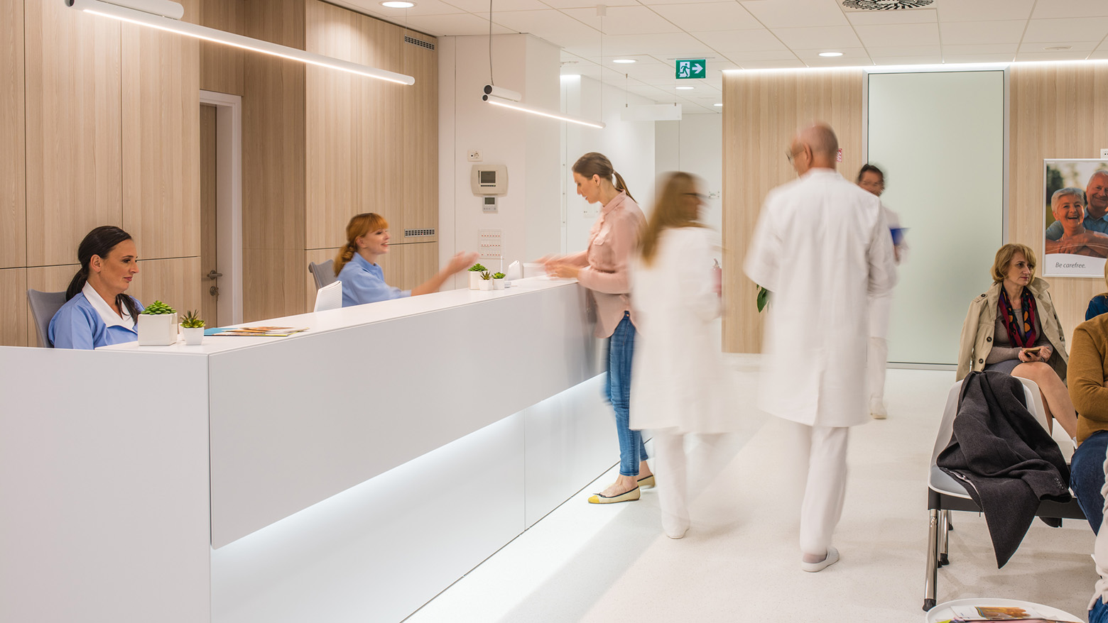 A clinic waiting room with seating patients, doctors walking by, and receptionists at a desk