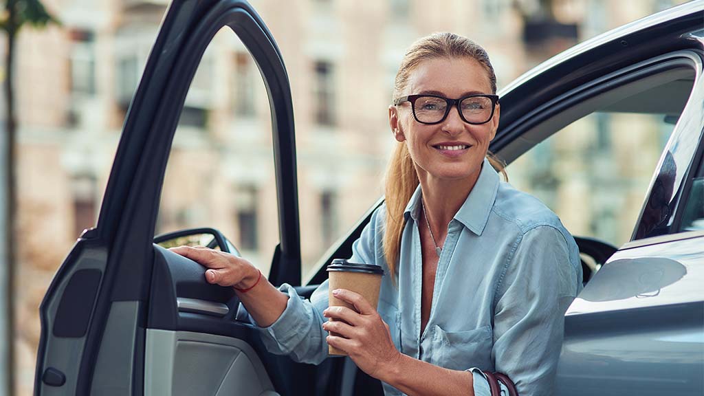 A woman steps out of the driver's seat of her car while holding a cup of coffee
