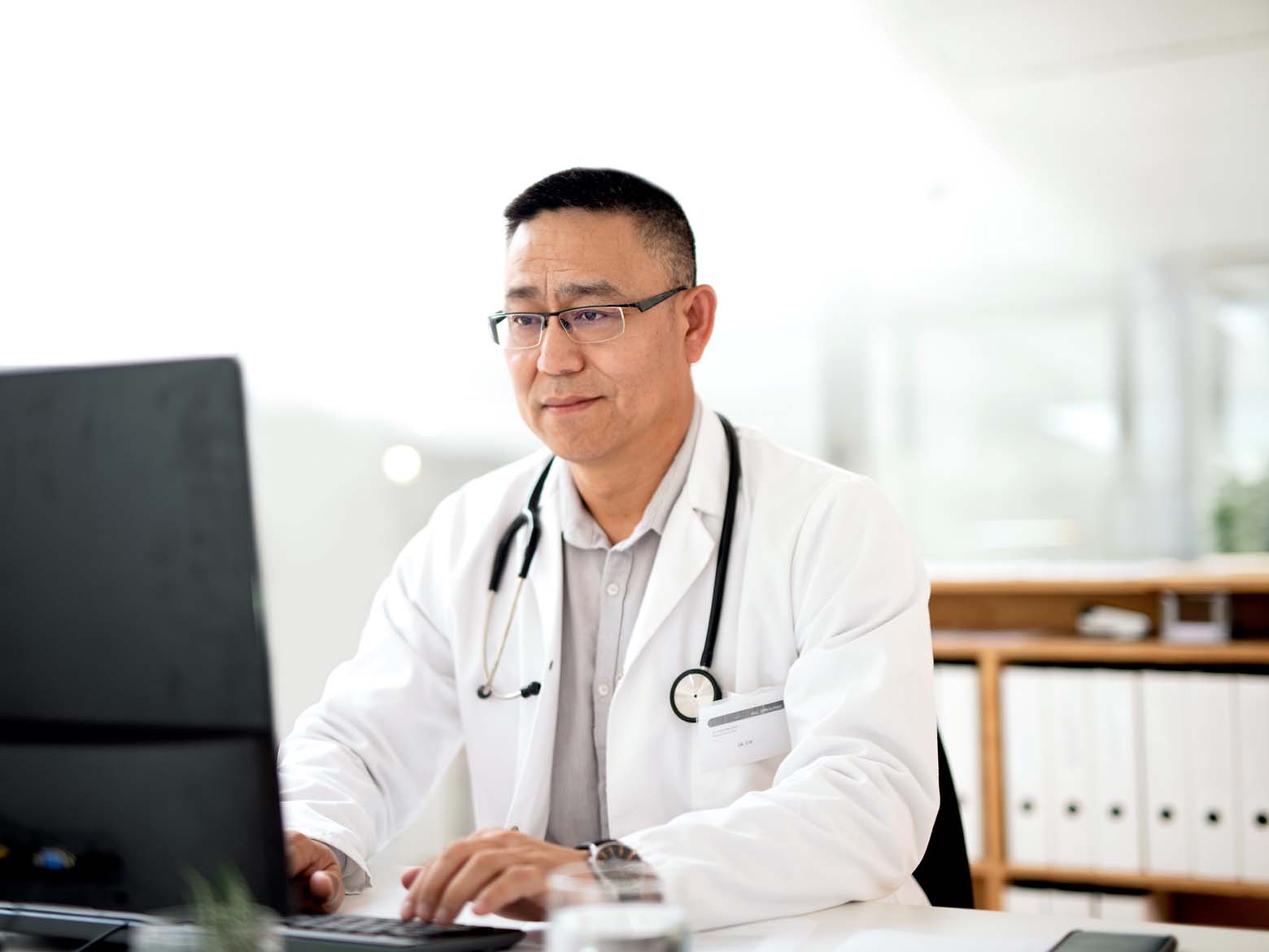 A male doctor sits at a desk while using a desktop computer