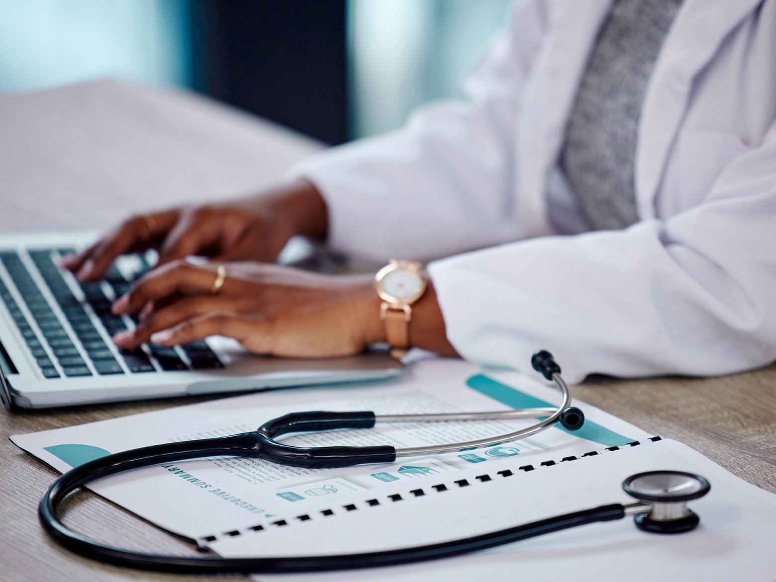 A doctor's hands typing on a laptop next to a stethoscope