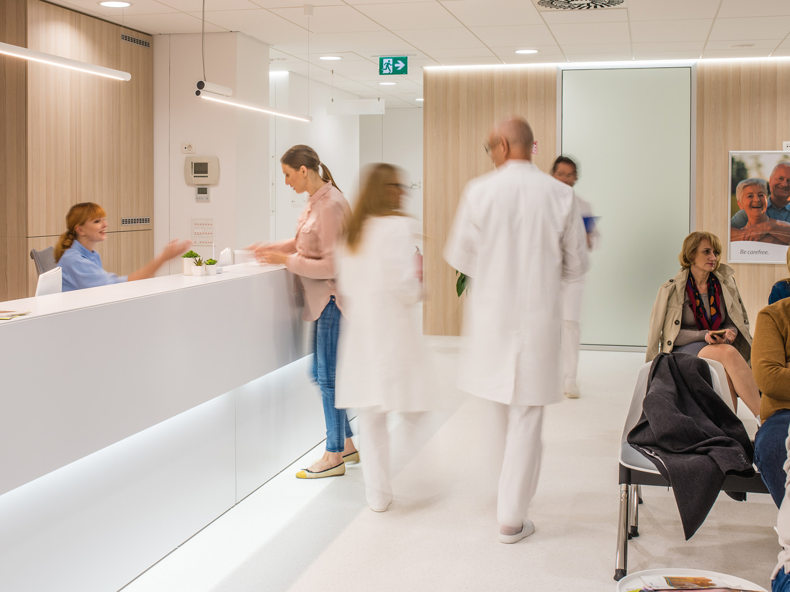 A clinic waiting room with seating patients, doctors walking by, and receptionists at a desk