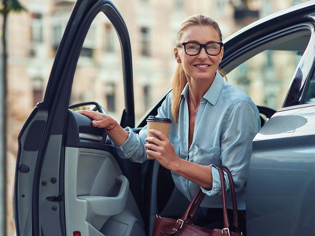A woman steps out of the driver's seat of her car while holding a cup of coffee